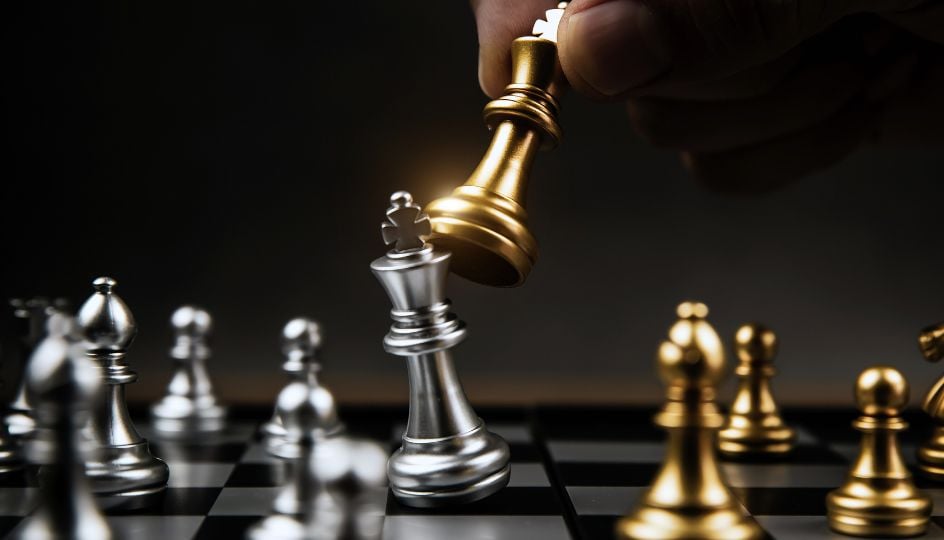 Close-up of a chessboard where a hand lifts a gold king piece above a silver king, surrounded by other gold and silver chess pieces, symbolizing a decisive move or checkmate under dramatic lighting.